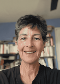 Smiling woman with short dark hair wearing a black shirt, indoors with a blurred background of bookshelves.