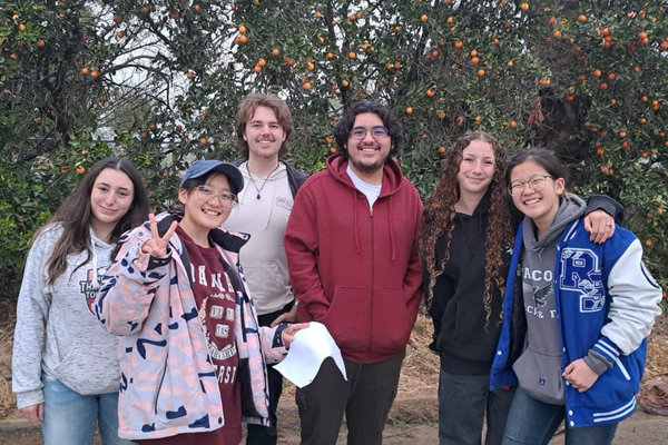 A group of smiling high school students pose together in front of orange trees in an orchard, holding papers and wearing casual clothing.