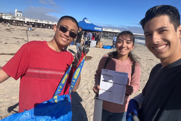 Three smiling students hold beach cleanup tools, a clipboard, and a trash bag during a volunteer event on the sand near a pier.