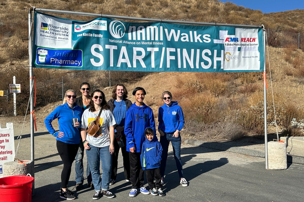 A group of participants pose under a banner reading “NAMIWalks START/FINISH” at a mental health awareness walk in a dry, hilly outdoor area.