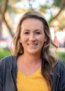 Portrait of a woman smiling outdoors, wearing a yellow shirt and gray hoodie.