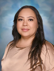 Professional headshot of Belen De La Cruz, wearing a beige blouse and smiling in front of a soft blue backdrop.