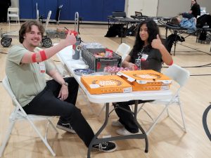 Two smiling individuals with red bandages on their arms give a thumbs-up while seated at a snack table in a gymnasium after donating blood. The table has pizza boxes, snacks, and water bottles, with blood donation stations visible in the background.