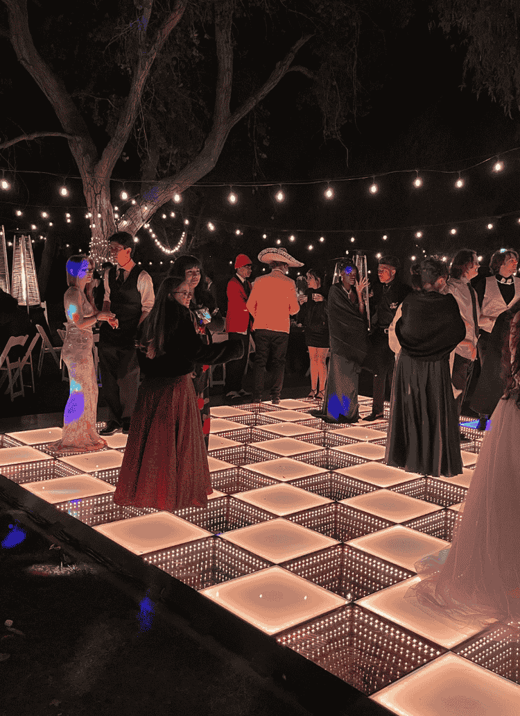 Students in formal attire enjoy dancing and mingling on a glowing LED dance floor under string lights during prom night.