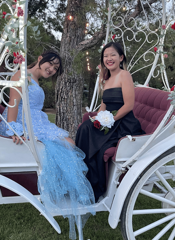 Two students dressed in formal prom gowns smile while seated in a decorative white carriage adorned with flowers and string lights.