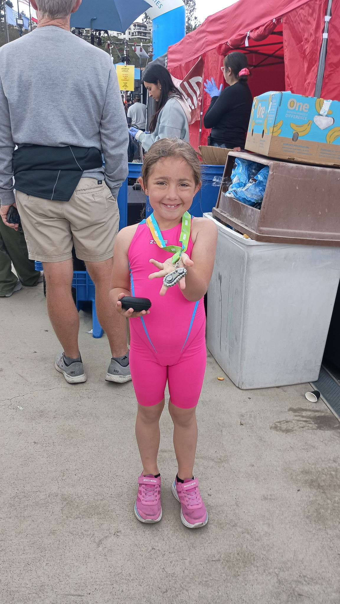 Young girl in a pink athletic suit smiles while showing her medal after completing an event, standing in front of a booth with volunteers.