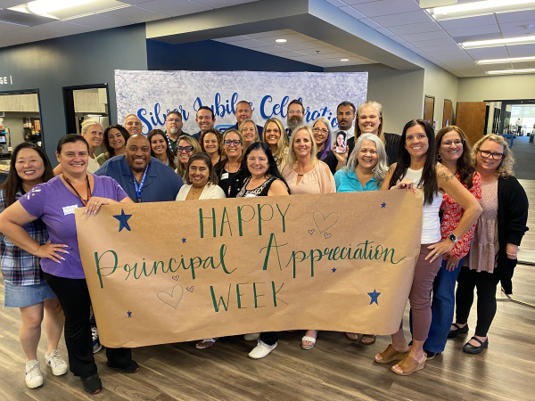 A group of staff members smile together in front of a handmade sign that reads “Happy Principal Appreciation Week.”