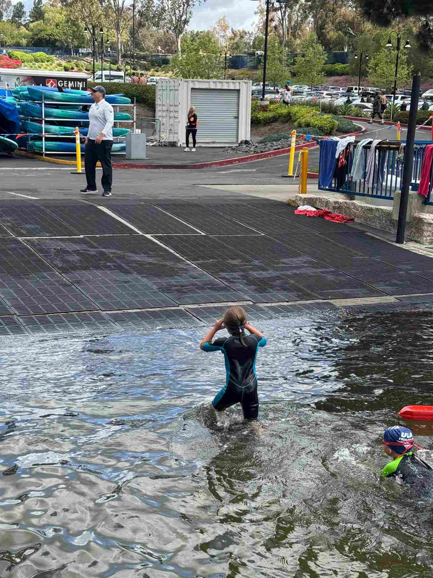 Two students in wetsuits participate in a water activity at a launch ramp, while an instructor observes from the shore.