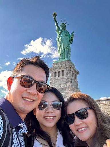 A family smiles for a selfie in front of the Statue of Liberty on a sunny day, all wearing sunglasses.