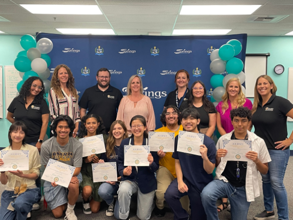 A group of high school seniors pose proudly with certificates in front of a Springs Charter Schools backdrop during Senior Signing Day, joined by staff and teachers.
