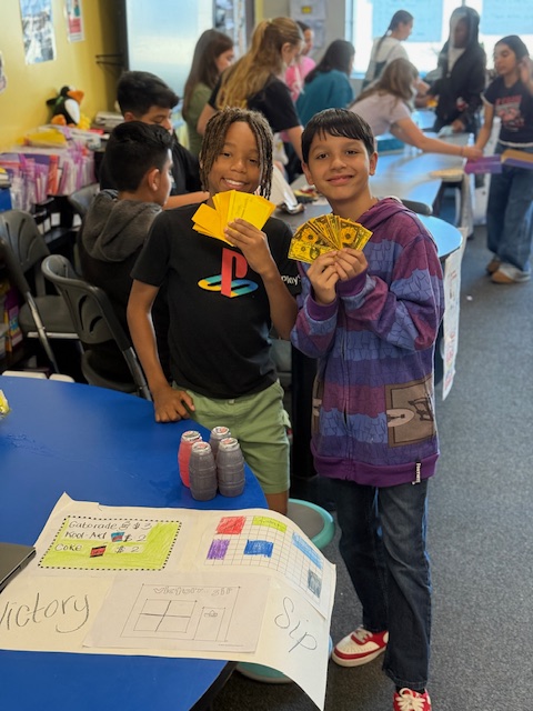 Two smiling students proudly hold play money behind their business booth, “Victory Sip,” during a classroom marketplace event.