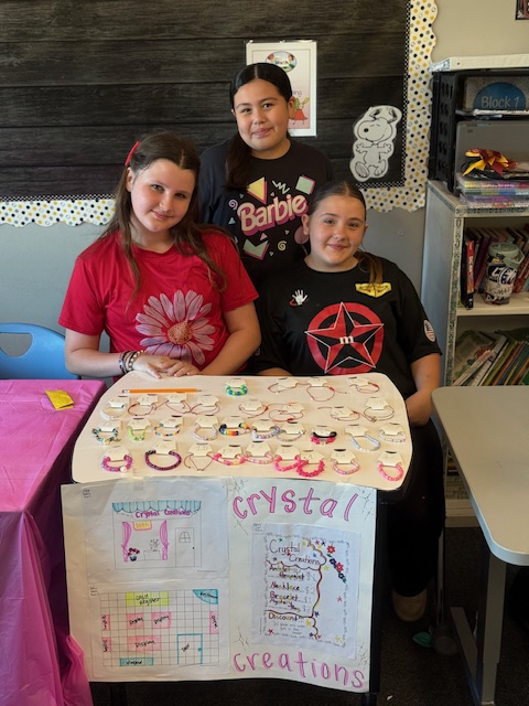 Three students smile behind a display of handmade bracelets at their classroom business booth labeled “Crystal Creations.”