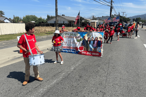 Students from Cherry Valley Student Center marching in a street parade, holding a Springs Charter Schools banner and playing drums.
