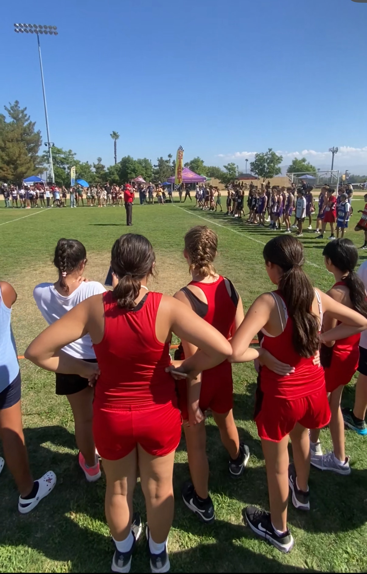 Students in athletic uniforms standing on a grassy field preparing for a cross country race with other teams lined up in the distance.