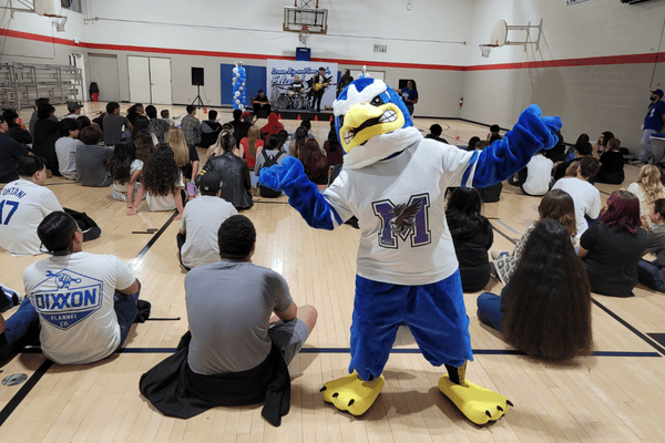 School mascot posing in front of students seated in a gymnasium assembly.