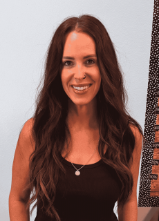 Woman with long brown hair and a nose ring smiling in front of a light blue wall.