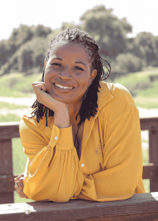 Woman smiling and leaning on a wooden railing outdoors, wearing a bright yellow blouse.