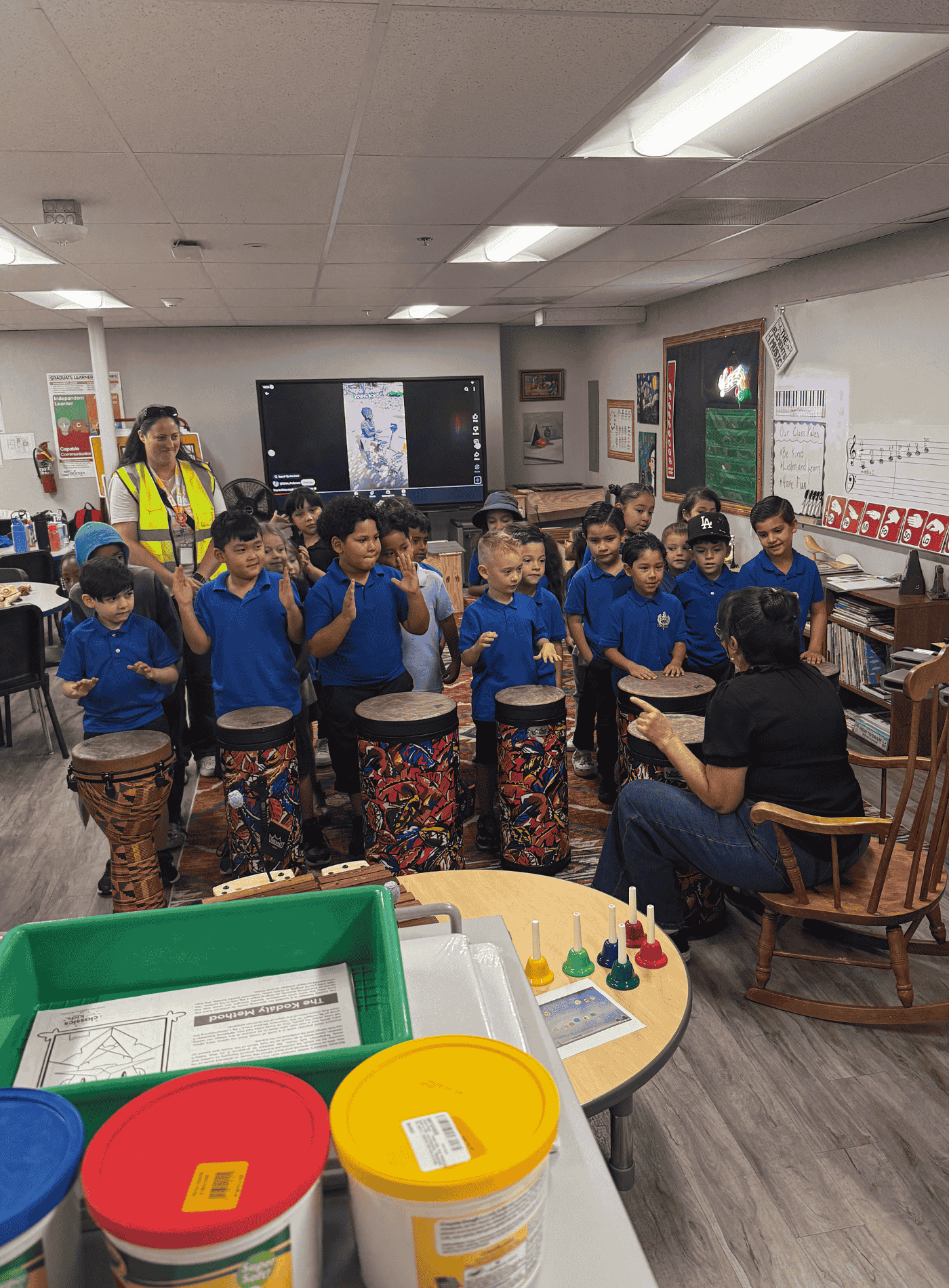 Elementary students in blue uniforms participate in a drumming lesson with their teacher in a music classroom.