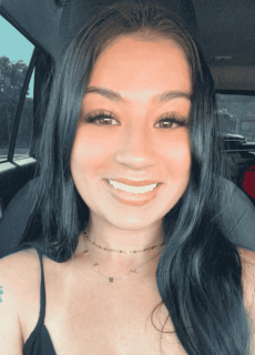 Smiling woman with long dark hair, wearing a black top and gold necklaces, seated in a car.