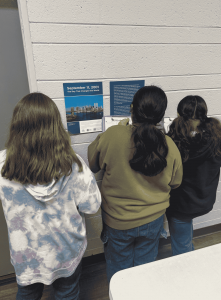 Three students stand in front of a September 11, 2001 educational display, taking notes on clipboards.
