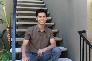 Young man smiling while seated on outdoor steps, wearing a brown polo shirt and jeans.