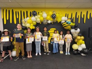 Springs Charter Schools spelling bee winners stand on stage holding medals and ribbons under a bee-themed balloon arch.