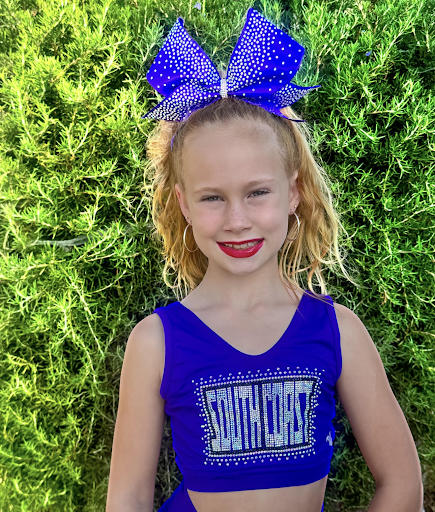 Springs Charter Schools student wearing a blue cheer outfit and bow, smiling outdoors in front of greenery.