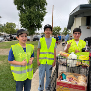 Students wearing safety vests standing next to a cart filled with food items during a community service activity.