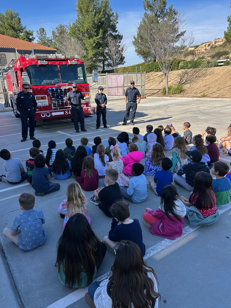 Firefighters speaking to a group of students seated on campus in front of a fire truck.