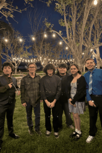 Students dressed formally posing together during a school dance event under string lights outdoors.
