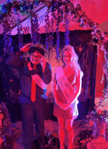 Students wearing crowns and sashes posing at a decorated school dance photo backdrop.