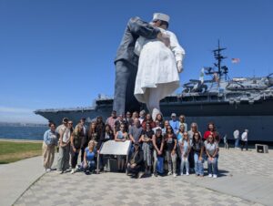 Group of people posing in front of a large naval statue and ship by the waterfront.