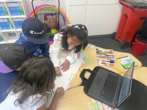Three students working together at a table drawing on paper with markers and pencils.