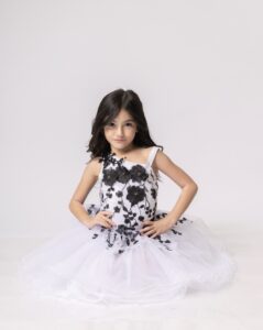 Young student posing in a white dress with black floral details against a plain background.