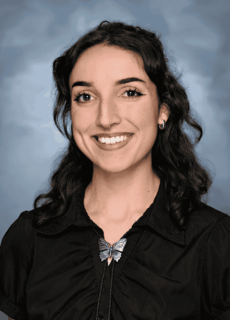 Smiling woman with dark wavy hair wearing a black blouse and butterfly necklace against a neutral background.