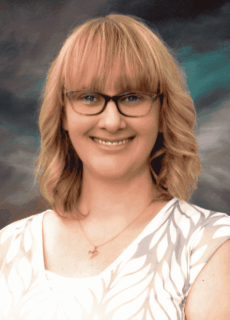 Smiling woman with blonde shoulder-length hair and glasses wearing a light patterned top.