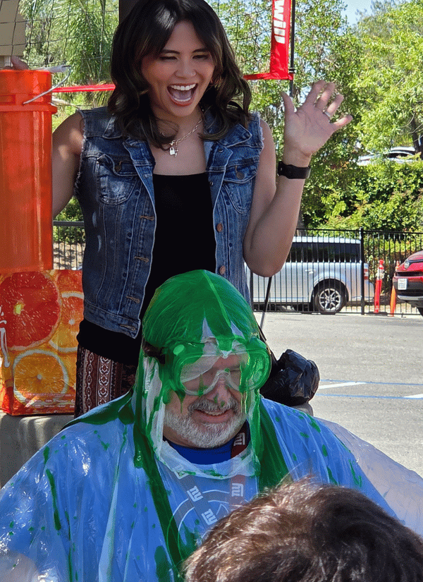 A smiling woman in a denim vest cheering as green slime is poured over a man wearing safety goggles and a clear plastic poncho outdoors.