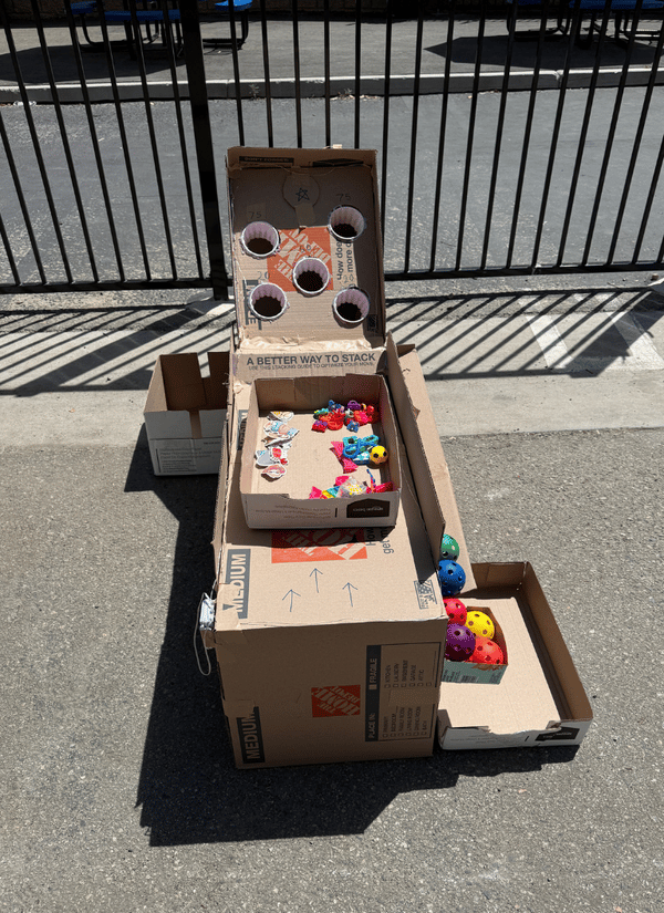 A handmade cardboard skee-ball style game crafted from recycled boxes, paper cups, and plastic colorful balls set up outdoors on a paved area.