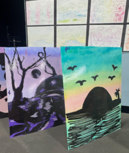 Two colorful student paintings of landscapes displayed on a table in a classroom.