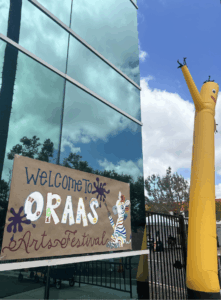 Welcome sign for the ORAA Arts Festival displayed beside a yellow inflatable dancer outside a school building.