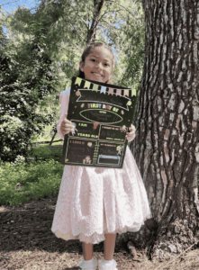 Young student standing outdoors beside a tree while holding a “First Day of School” sign with personal goals and favorite activities written on it.