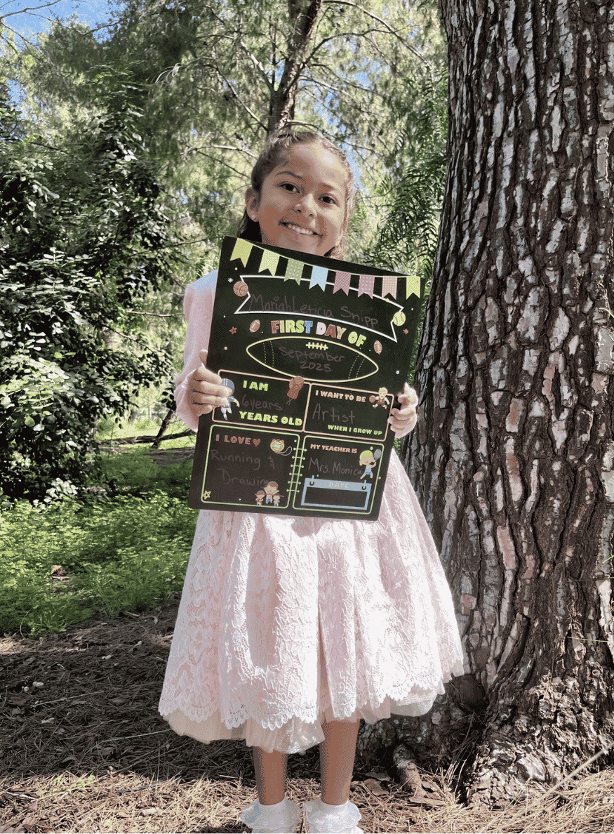 Young student standing outdoors beside a tree while holding a “First Day of School” sign with personal goals and favorite activities written on it.