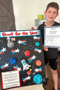 Elementary student holding a certificate beside a space-themed “Shoot for the Stars” school project display.