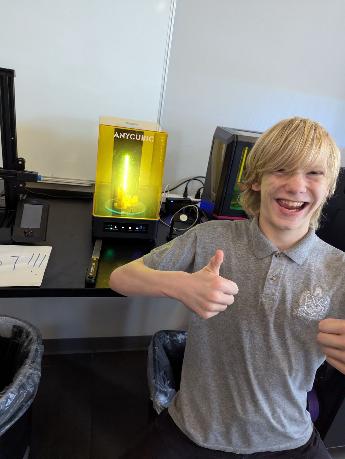 A smiling male student in a grey school polo shirt giving two thumbs up in front of an illuminated UV resin 3D print curing station.