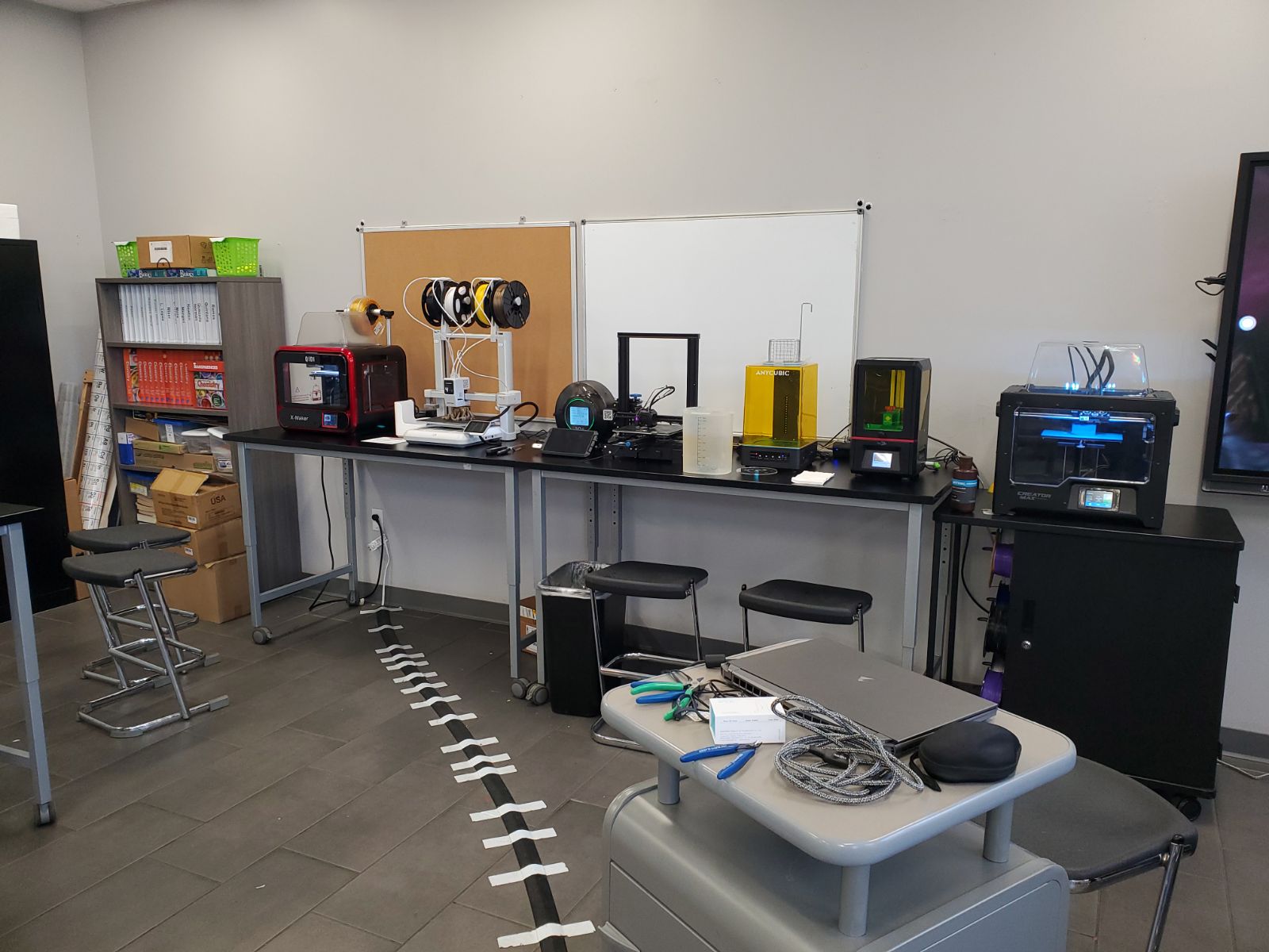 A row of various 3D printers lined up on lab tables in a classroom, featuring filament spools, resin curing stations, safety tools, and a laptop setup.