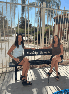 Principal Valerie Walker and Vice Principal Jeanette LaRue sitting on a black metal Buddy Bench outdoors in front of a white fenced area.