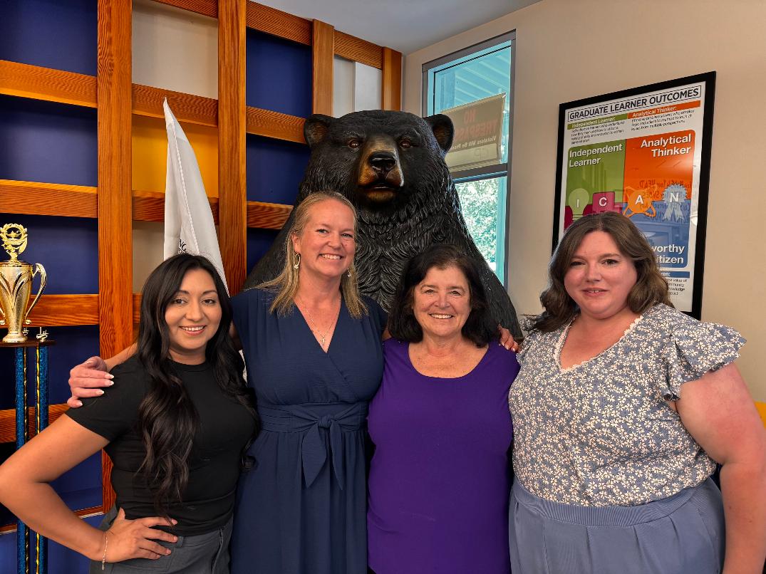 Four Bear River Student Center staff members smiling together indoors in front of a large black bear statue and a Graduate Learner Outcomes poster.