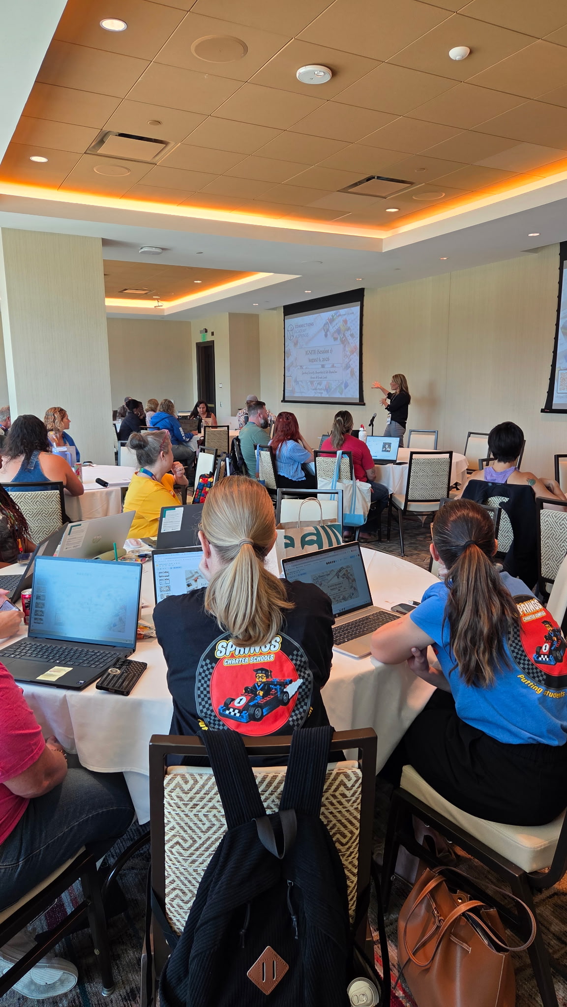 Participants seated at round tables with open laptops facing a presentation screen during a professional IGNITE session.