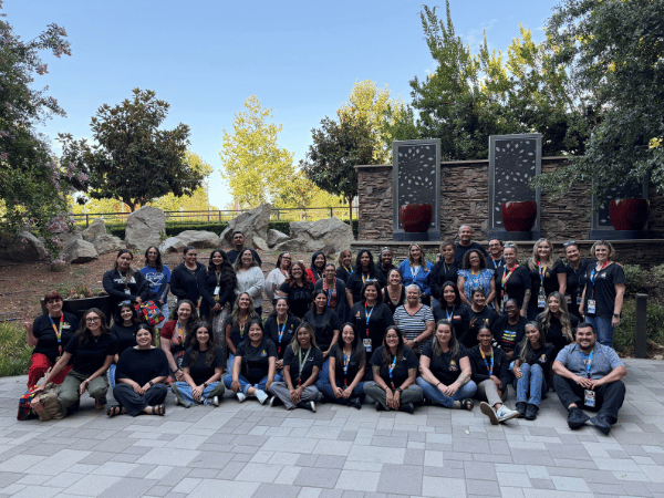 A large group of staff members wearing lanyards gathered together for a group photo outdoors on a stone patio in front of a decorative fountain and rock wall landscape.