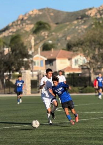 High school students actively pursuing the ball during a competitive outdoor soccer match on a green field with hills in the background.